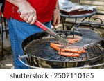 Hot dogs on charcoal kettle grill being turned by older man with tongs