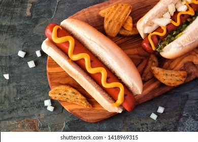Hot dogs with toppings and potato wedges on wooden board. Close up, top view.