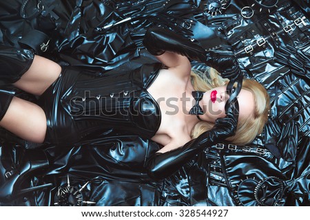 Hot dominant beautiful blonde mistress woman in shiny latex fetish corset and mask posing on bdsm accessories and fetish clothing