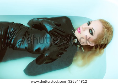Hot dominant blonde mistress woman in wet shiny latex fetish dress, gloves and spiky leather collar posing in white bath with blue water