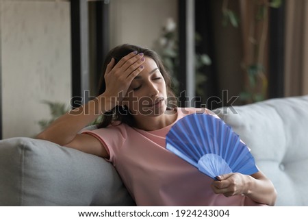 Too hot. Exhausted indian female suffer from heat in modern apartment use hand fan feel lack of air conditioning. Unhappy young mixed race female complaining on high temperature at home hard to breath