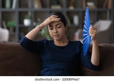 So Hot. Frustrated Young Indian Lady Sit On Sofa Suffer From Heat At Living Room Touch Forehead Feel Migraine Heatstroke. Sad Unhealthy Teen Female Breath Warm Dry Air Try To Cool Herself By Paper Fan