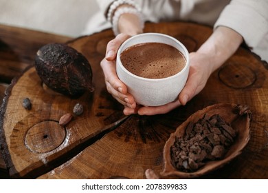Cacao ceremonial caliente hecho a mano en taza blanca. Manos de mujer sosteniendo cacao artesanal, vista superior sobre una mesa de madera. Bebé de chocolate orgánico y saludable preparado con frijoles, sin azúcar. Ceremonia de entrega, acogedor café