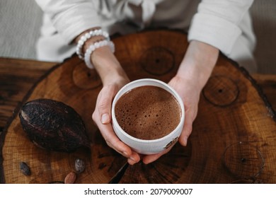 Hot handmade ceremonial cacao in white cup. Woman hands holding craft cocoa, top view on wooden table. Organic healthy chocolate drink prepared from beans, no sugar. Giving cup on ceremony, cozy cafe