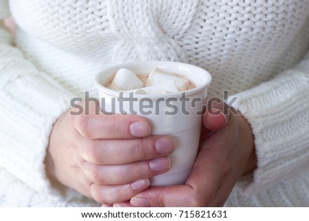 Hot homemade cocoa with marshmallow in the hands of a woman. Woman in a white sweater. Rustic style, selective focus.
