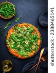 Hot homemade pizza with blue cheese, ham, pesto sauce and fresh arugula, black table background, top view