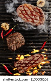 Hot Juicy Chivas Beef Steak On Barbecue Flames Grill Background. Smoked Ribeye Beef Steaks With Grilled Garlic, Spices And Red Peppers. Bbq Entrecote Meat Top View