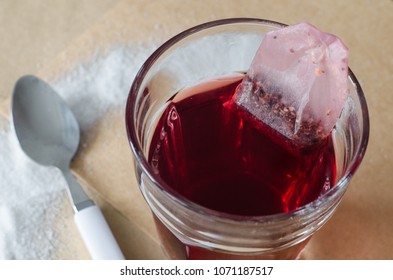 Hot Red Berry Tea And Tea Bag In Glass, Spilled Sugar Around And White Spoon Near, On Bright Brown Paper Background