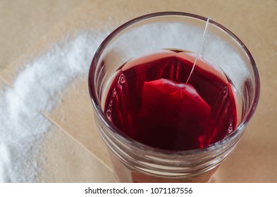 Hot Red Tea And Tea Bag In Glass, Spilled Sugar Around On Bright Brown Paper Background