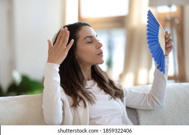 Too Hot In The Room. Stressed Sad Millennial Female Sitting On Couch At Home Waving Herself With Hand Fan Suffering From Extra Heat, Feeling Tired Overheated Exhausted Hard To Breath With Warm Dry Air