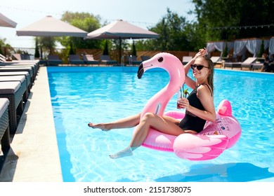 Hot Slim Woman In Bikini Chilling With Cocktail On Inflatable Pink Flamingo Float At Swimming Pool. Fit Girl In Swimwear Lies In The Sun And Drinks On Floaty. Pretty Female On Tropical Vacation.