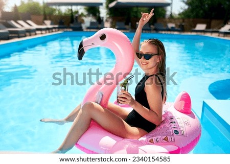 Hot slim woman in bikini chilling with cocktail on inflatable pink flamingo float at swimming pool. Fit girl in swimwear lies in the sun and drinks on floaty. Pretty female on tropical vacation.