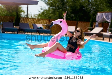 Hot slim woman have fun and wave hand on inflatable pink flamingo float mattress in bikini at swimming pool. Attractive fit girl in swimwear lies in sun on floaty. Pretty female on tropical vacation.
