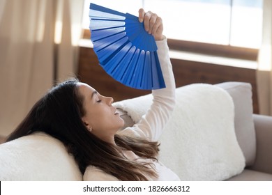 Too Hot Summer Day. Exhausted Prostrated Female Resting On Couch At Living Room Fanning Herself With Hand Waver Suffering From Heat Dreaming About Conditioner System At Home To Breath Cool Fresh Air