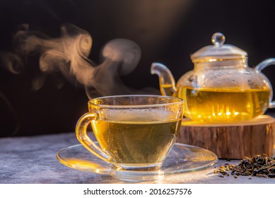 Hot tea in glass teapot and cup with steam on wooden table and black background