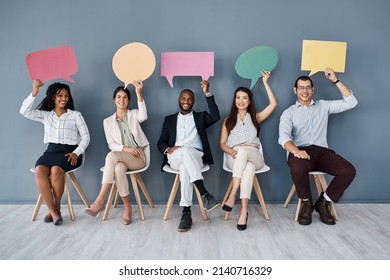 Its A Hot Topic In The Business World Right Now. Portrait Of A Group Of Businesspeople Holding Speech Bubbles While Sitting In Line Against A Grey Background.