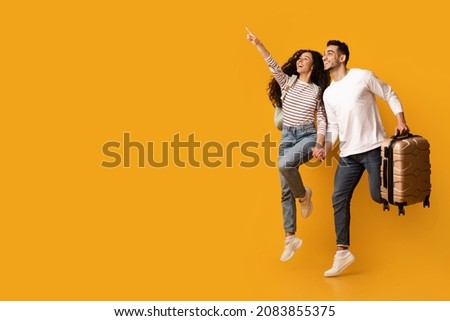Hot Tours. Joyful Arab Couple With Suitcase Jumping And Pointing Away, Excited Young Middle Eastern Spouses Showing Copy Space On Yellow Background For Travel Offer Or Advertisement, Full Length