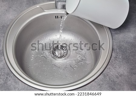 Hot water pours from a white electric kettle into a metal kitchen sink. Debris cleaning and disinfection.