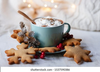 Hot winter drink: cappuccino with whipped cream in blue mug. Christmas time. Cozy home atmosphere, white background, copy space for text. Homemade gingerbread cookies, cones, christmas lights