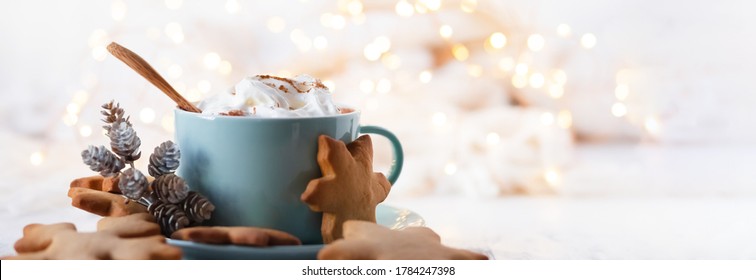 Hot winter drink: chocolate with whipped cream in blue mug. Christmas time. Cozy home atmosphere, white background. Homemade gingerbread cookies, christmas lights. Banner, copy space for text