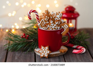 Hot winter drink in a red mug: cozy home composition with gingerbread cookies, candy cane, fir tree branch. Wooden background, christmas lights and candles - Powered by Shutterstock