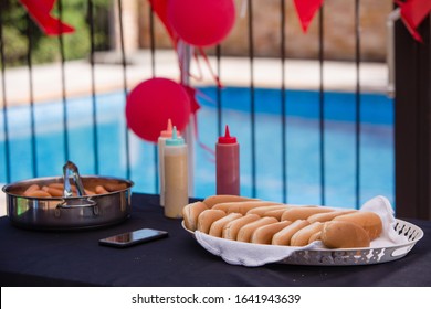 Hot-dogs And Hot-dog Bread Buns On Birthday Celebration Table By The Pool. Smart Phone Left By The Food, Ketchup, Mustard, And Mayonnaise Squeeze Bottles. House Party Decorations.