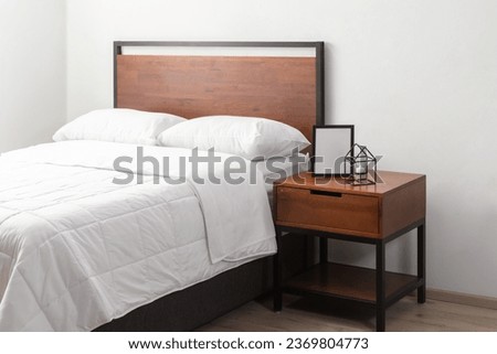 Hotel Bedroom Interior Featuring a King-Size Metal Wood Platform Bed with Headboard, White Cozy Bed Blanket, and a Bedside Table Adorned with a Decorative Ornaments, Set Against a White Wall.