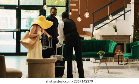 Hotel bellhop providing luxury service to guests entering reception lobby, concierge carrying baggage and trolley bags to room. Young couple arriving at tropical resort front desk. - Powered by Shutterstock