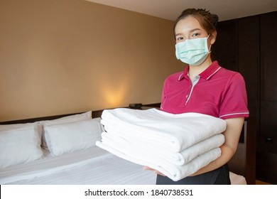 Hotel Maid Wearing A Mask Is Looking At Camera While Holding A Stack Of Fresh Towels With Twin Bedroom In The Background.