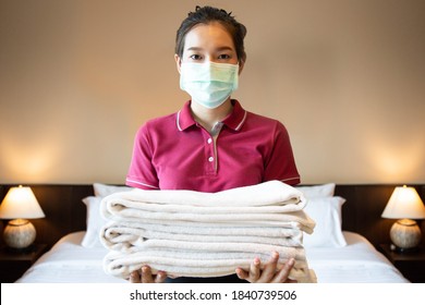 Hotel Maid Wearing A Mask Is Looking At Camera While Holding A Stack Of Fresh Towels With Twin Bedroom In The Background. Front View Shot.