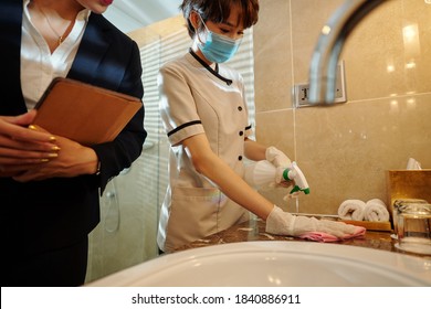 Hotel Manager Controlling Hotel Maid In Medical Mask Wiping Surfaces In Bathroom With Disinfecting Spray