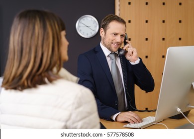 Hotel Manager Taking A Booking Watched By A Female Client As He Checks Availability On The Computer