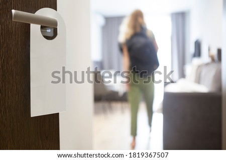 The hotel room with empty please do not disturb sign on the door with blurred person in room. Young woman with backpack. 