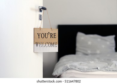 Hotel Room, You Are Welcome Words On Wooden Vintage Frame Card Background, Hanging On The Bedroom Door, Enter The Home. Welcoming Hotel Bed With Pillows And Backlight From The Window. 