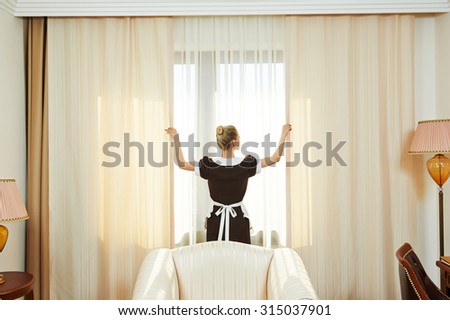 Hotel service. female housekeeping chambermaid worker with opening curtains of window in room