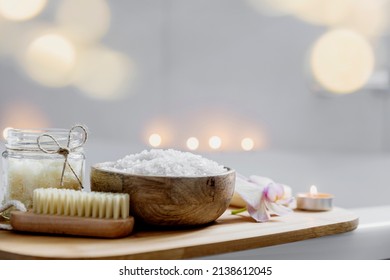 Hotel spa treatment, home bath procedure. White washbasin in bathroom, accessories on tray. Burning candles, soap, foot brush, towel, glass bottle with sea salt, orchid flower. Blurred background