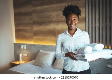 Hotel staff setting up pillow on bed. African housekeeper looking at the camera holding a towel a hotel room. Clean towels during housekeeping in a hotel room