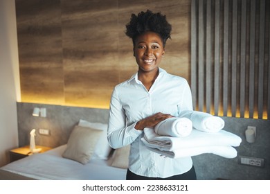 Hotel staff setting up pillow on bed. African housekeeper looking at the camera holding a towel a hotel room. Clean towels during housekeeping in a hotel room - Shutterstock ID 2238633121