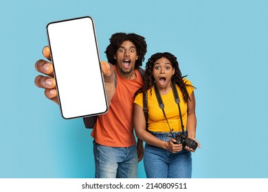 Hotel And Tickets Online Booking Concept. Amazed Young Black Couple With Camera And Backpack Tourists Showing Modern Smartphone With White Empty Screen, Mockup, Blue Studio Background, Copy Space