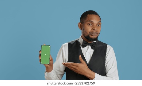 Hotel worker bellhop points at phone with isolated mockup display, showing green screen against blue background in studio. Hotelier doorman greeting resort guests at reception. Camera A. - Powered by Shutterstock