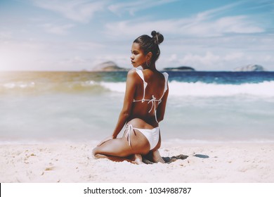 Hottie young African American female model is sittingÂ on the sand half-turned to camera in front of ocean; rear view of foxy Brazilian girl sitting on the beach of warm summer sea with horizon behind