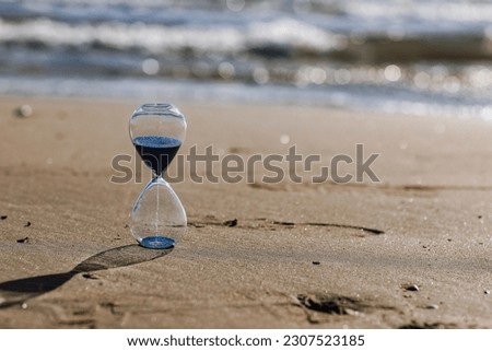 Hourglass with blue sand inside in a warm golden morning sunlight with shadow on a sandy beach and sea background, starting time for a new day or running of time. Concept of the rapid passage of time.