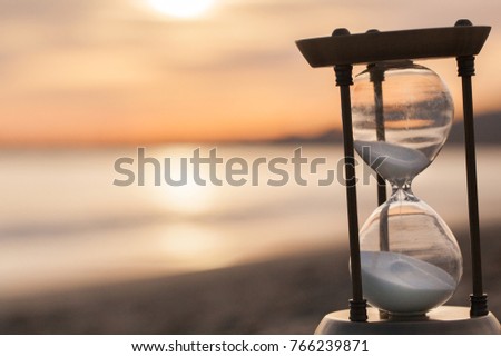 Hourglass in the dawn time. Sand passing through the glass bulbs of an hourglass measuring the passing time as it counts down to a deadline or closure on a sunset/ sunrise beach background. 