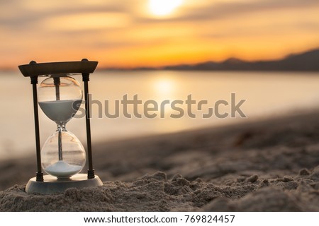 Hourglass in the sunset golden hour. Sand passing through the glass bulbs of an hourglass measuring the passing time as it counts down to a deadline or closure on a sunset/ sunrise beach background. 