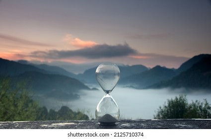 A hourglass (with falling sand) on a wooden wet table with sea of fog and mountain range silhouette in sunrise time - Shutterstock ID 2125710374