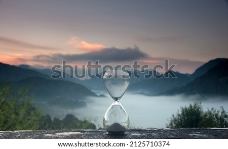 A hourglass (with falling sand) on a wooden wet table with sea of fog and mountain range silhouette in sunrise time