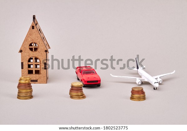 House, car and\
airplane cost concept. Models of house car and airplane with stacks\
of coins isolated on\
grey.