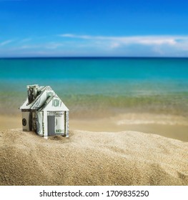 House Carved Out Of Dollar Bills On The Sea Sand. Concept On The Topic Of Selling Or Buying A House, Buying A House On The Sea Or Ocean Shore