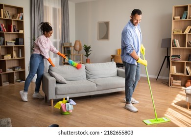 House Chores. African American Husband And Wife Cleaning Modern Living Room With Mop And Feather Duster Standing At Home On Weekend. Spouses Doing Housework Together