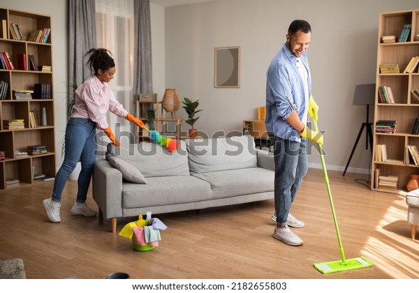 House Chores. African\
American Husband And Wife Cleaning Modern Living Room With Mop And\
Feather Duster Standing At Home On Weekend. Spouses Doing Housework\
Together